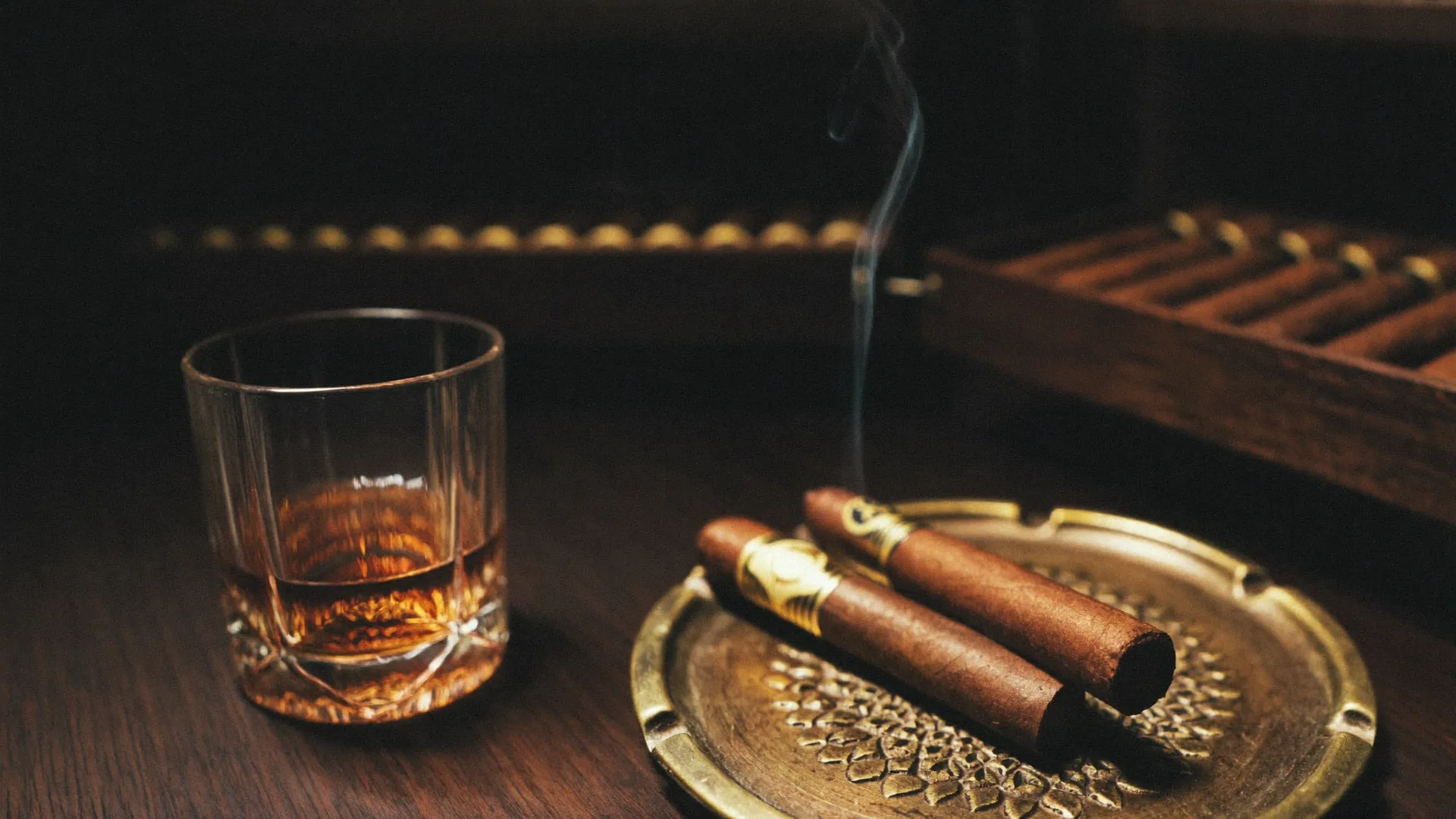 Two premium cigars on an aged brass ashtray beside a glass of amber whisky, a backlit humidor behind